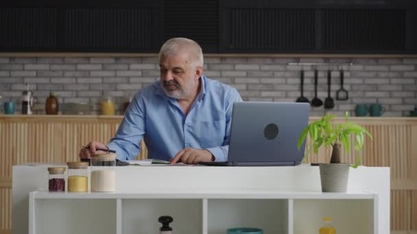 Старик проверяет коммунальные счета, рассчитывает и оплачивает онлайн, используя ноутбук, сидя дома на кухне — стоковое видео