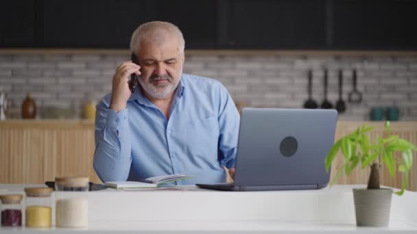 Homme d'affaires âgé communique par téléphone, travaille à la maison, assis à la cuisine avec ordinateur portable et papiers sur la table — Video