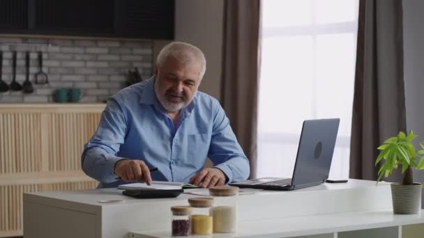 Hombre viejo está trabajando en casa, haciendo cálculos y buscando información en el ordenador portátil, sentado solo en la cocina, trabajo a distancia — Vídeos de Stock