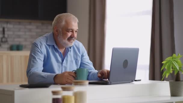 Пожилой человек серфинг интернет на ноутбуке, отправка сообщения в онлайн чате, сидя в одиночестве на кухне и пить чай — стоковое видео