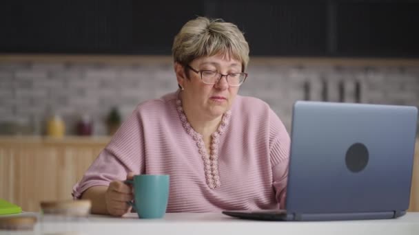 Mujer jubilada está pasando tiempo en Internet, utilizando el ordenador portátil sentado en la cocina de casa durante el día, retrato de la anciana con portátil moderno — Vídeos de Stock