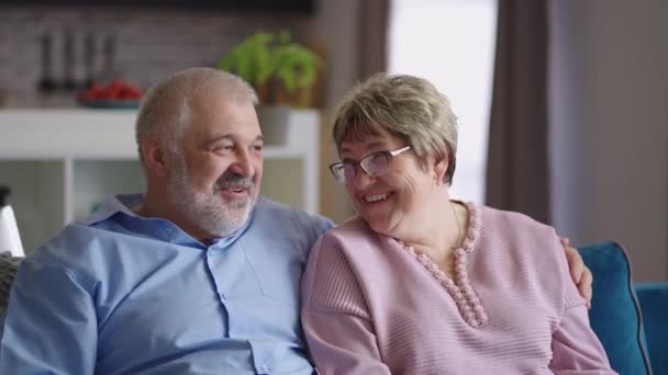 Пожилая супружеская пара отдыхает в гостиной, старик и женщина общаются и смеются, портрет снят в помещении — стоковое видео