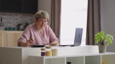 Yaşlı kadın ev mutfağında dizüstü bilgisayarla çalışıyor. Evrakları kontrol ediyor ve programı dolduruyor.