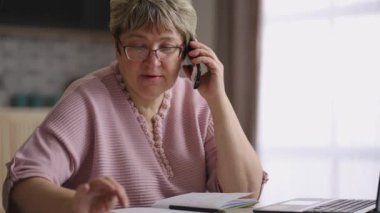 Gözlüklü yaşlı kadın evde çalışıyor, dizüstü bilgisayar kullanıyor ve telefonla arıyor, hesap makinesiyle sayıyor.