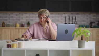 Orta yaşlı kadın evden uzaktan çalışıyor, iş arkadaşlarıyla telefonla iletişim kuruyor, hesap makinesi ve dizüstü bilgisayar kullanarak rapor hazırlıyor