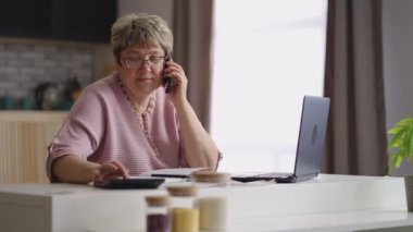 Yaşlı kadın telefonla arıyor, evden çalışıyor, mutfağında tek başına oturuyor, dizüstü bilgisayar ve hesap makinesi kullanıyor.