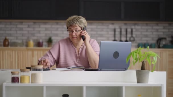 Donna di mezza età sta lavorando in remoto da casa, comunicare con i colleghi per telefono, utilizzando calcolatrice e laptop per fare rapporto — Video Stock
