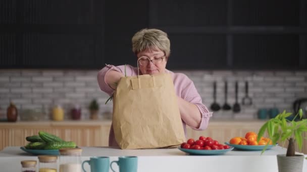 Vecchia donna sta disimballando il cibo dopo lo shopping in drogheria, mettendo verde fresco e baguette sul tavolo della cucina — Video Stock