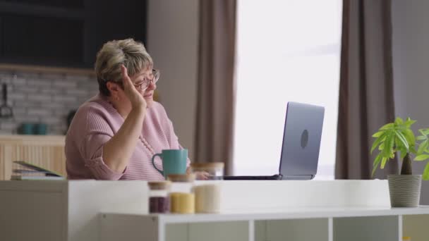 Ηλικιωμένη κυρία συνομιλεί online με την οικογένεια με βιντεοκλήση στο laptop, ηλικιωμένη γυναίκα κάθεται στο σπίτι κουζίνα το Σαββατοκύριακο — Αρχείο Βίντεο