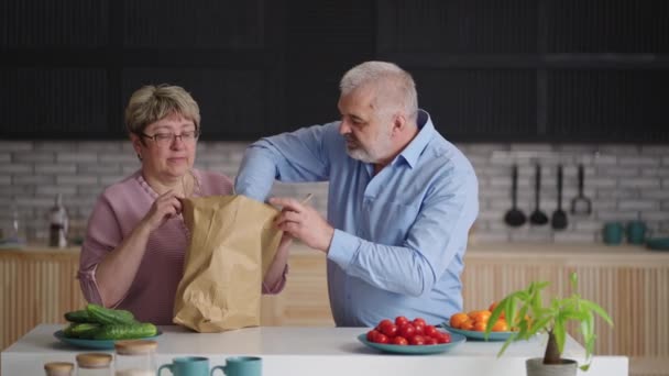 Rentner packen Tüte mit frischen Lebensmitteln in der Küche aus, Lieferung von Lebensmitteln nach Hause, alte Ehepartner legen Grün und Brot auf den Küchentisch — Stockvideo