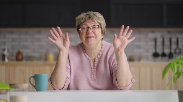 Mujer de edad linda está sentado en la cocina y mirando a la cámara, hablando y agitando las manos para dar la bienvenida, concepto de video chat en línea — Vídeo de stock