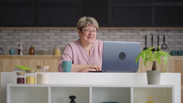 Fröhliche alte Frau kommuniziert per Videochat im Laptop, Rentnerin sitzt am Wochenende in der heimischen Küche — Stockvideo