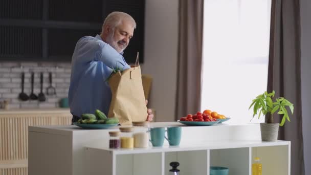 Hombre de pelo gris está desempacando la comida después de comprar en la tienda de comestibles, poniendo vegetación fresca y baguette en la mesa de la cocina — Vídeos de Stock