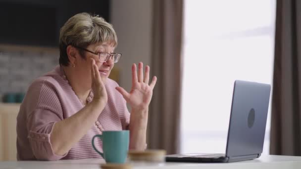 Senhora idosa alegre está se comunicando com a família por vídeo chat no laptop, mulher idosa está sentada em casa cozinha no fim de semana — Vídeo de Stock