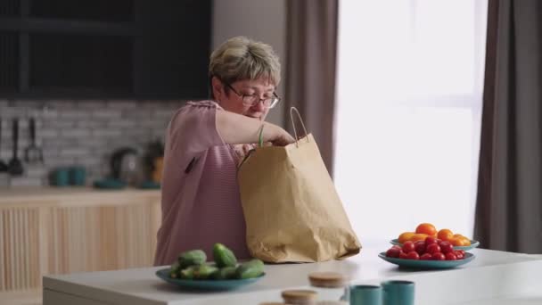 La abuela está desempacando la comida después de comprar en el mercado el fin de semana, tomando vegetación fresca y pan, retrato medio de una anciana soltera en casa — Vídeos de Stock