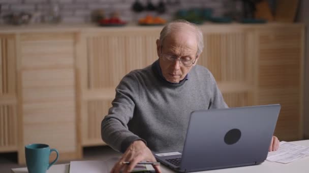 Az idős ember kiszámítja a kiadásait és a tervezett költségvetését, otthon ül, számológéppel számol és gépel a laptopon — Stock videók