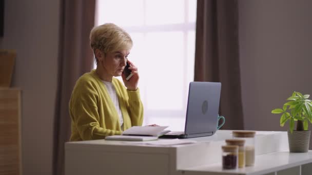 Vieja empresaria está trabajando en la oficina en casa, comunicándose por teléfono, sentado en la cocina y utilizando el ordenador portátil — Vídeos de Stock