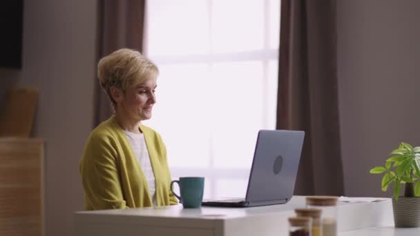 Gammal kvinna chattar med videosamtal i modern bärbar dator med internetuppkoppling sitter hemma kök, medium porträtt — Stockvideo