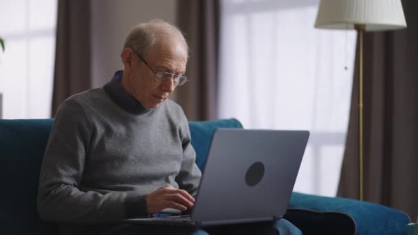 Åldrad man surfar internet med bärbar dator i sin lägenhet i helgen, pensionär skriver på tangentbord, kommunicera i sociala nät eller arbetar på distans — Stockvideo