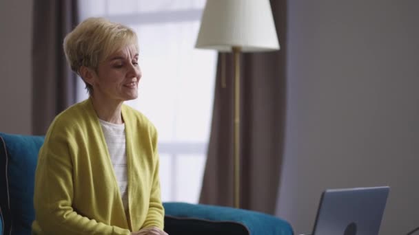 Mulher aposentada loira está sentada sozinha em casa e se comunicando por vídeo chat on-line com amigo, retrato de senhora idosa dentro de casa — Vídeo de Stock