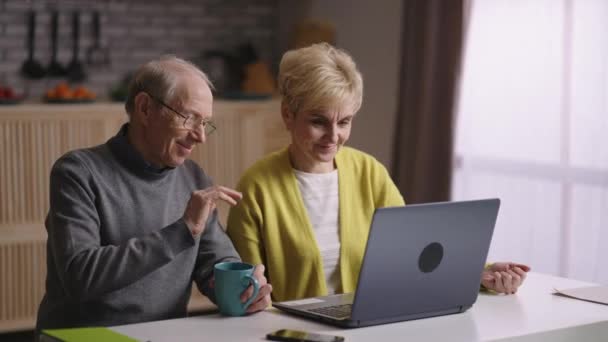 Vecchi sposi felici stanno chiacchierando tramite videochiamata con amici o familiari, sorridendo alla fotocamera web del computer portatile, seduti insieme in cucina — Video Stock