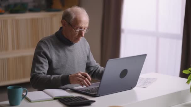 Anciano está trabajando a distancia, sentado en su oficina en casa, contando por calculadora y escribiendo en el portátil — Vídeos de Stock