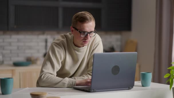 Seorang pria berkacamata bekerja dari rumah di dapurnya menggunakan laptop dan internet. Kerja jauh dan belajar jarak jauh — Stok Video