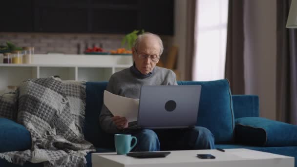 Ensam pensionerad man tittar på papper sitter i lägenheten, med laptop för att kontrollera, arbetar på distans hemifrån — Stockvideo
