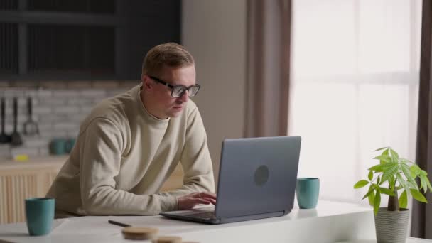 En man med glasögon arbetar hemifrån i sitt kök med hjälp av en laptop och Internet. Fjärrarbete och distansutbildning — Stockvideo
