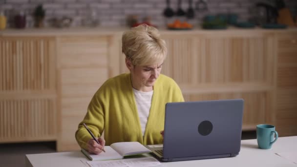 Autoeducación para los jubilados, mujer de edad avanzada está viendo tutorial en Internet por ordenador portátil y notas de escritura, sentado en casa — Vídeos de Stock