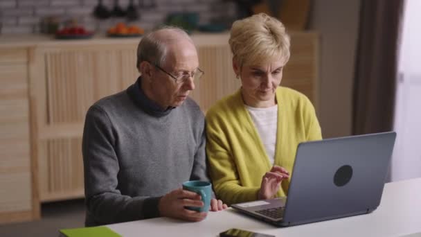 Casal idoso está usando laptop juntos, homem e mulher estão sentados em casa cozinha, compras on-line ou se comunicar em redes sociais — Vídeo de Stock