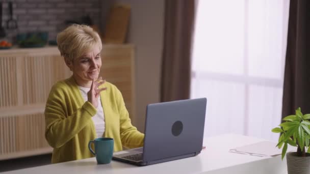 Счастливая бабушка звонит по видеосвязи в ноутбуке с друзьями или семьей, сидя одна на домашней кухне — стоковое видео