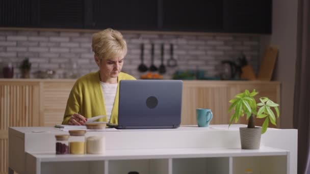 Старая учительница работает дома, проверяя домашнее задание учеников и учеников, сидя за столом на кухне — стоковое видео