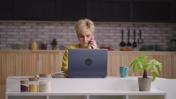 Äldre dam arbetar på distans, sitter hemma kök och kommunicera via mobiltelefon, med hjälp av laptop för att skicka e-post — Stockvideo