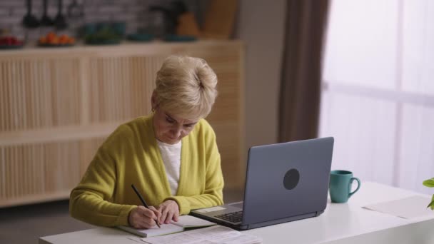 Old woman is learning online, sitting at home kitchen with laptop, looking for information in internet and making notes in notebook — Stock Video