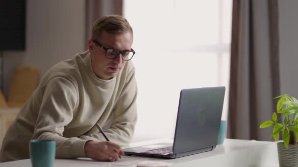 Cansado casualmente vestido homem estudando com computador sentado em casa. Jovem do sexo masculino com cabelo encaracolado ganhando conhecimento com a ajuda de dispositivo digital. — Vídeo de Stock