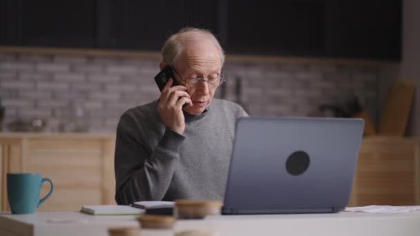 Γέρος εργάζεται με φορητό υπολογιστή στην κουζίνα στο σπίτι, καλώντας στο τηλέφωνο για συμβουλές, μέσο πορτρέτο των συνταξιούχων στο διαμέρισμα — Αρχείο Βίντεο