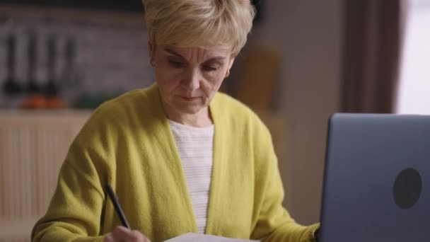 Oude dame is het leren van online of werken, surfen op internet in laptop en het maken van notities in notebook, portret van gepensioneerde vrouw thuis — Stockvideo