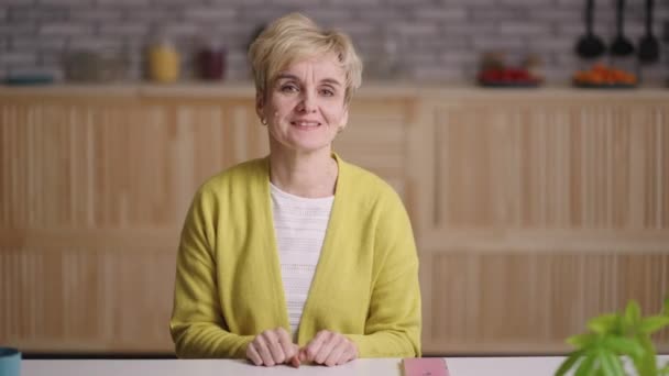 Ritratto di affascinante signora anziana guardando la macchina fotografica e agitando la mano per accogliere, seduto da solo in cucina — Video Stock