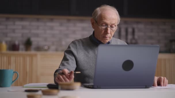 Старик подсчитывает коммунальные платежи и составляет бюджет, сидит один на кухне, работает с ноутбуком — стоковое видео