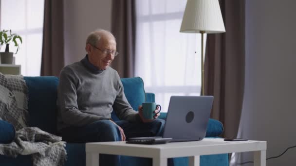 Man pensionär använder bärbar dator för att kommunicera via videochatt med vän eller familj, titta på webbkamera och dricka te — Stockvideo