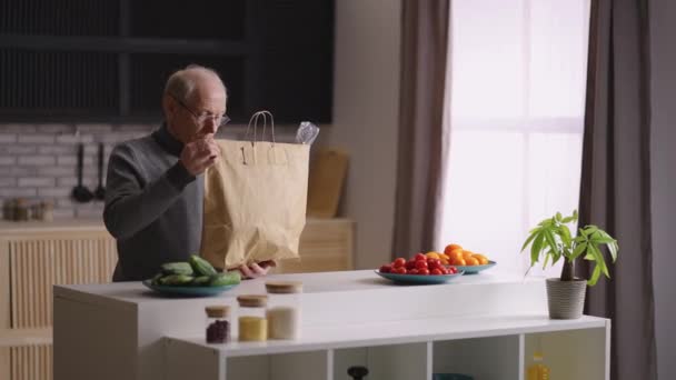 Hombre viejo soltero está desempacando la comida después de comprar en el mercado, poniendo mercancías en la mesa de la cocina — Vídeos de Stock