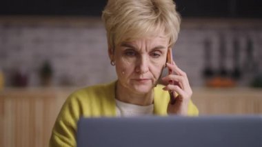 Yaşlı kadın uzaktan çalışıyor, evde oturuyor ve cep telefonuyla konuşuyor, dizüstü bilgisayarı kullanıyor, iş için interneti kullanıyor.