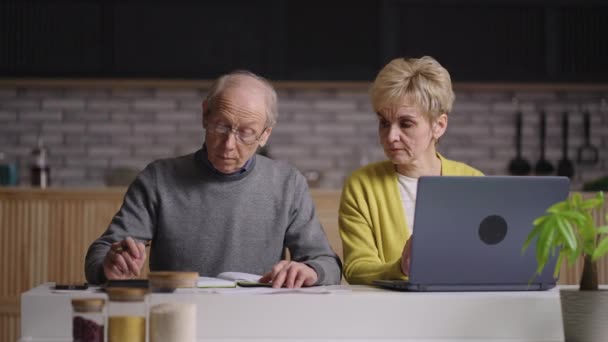 Vecchio e donna stanno pagando bollette di utilità online, seduti insieme a casa cucina, l'uomo sta calcolando e la donna sta utilizzando il computer portatile — Video Stock