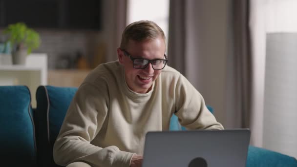 Homem sentado no sofá em casa fazendo chamada de vídeo usando computador portátil em auto-isolamento. Jovem professor loira feliz trabalhando em casa, falando em videoconferência laptop. — Vídeo de Stock