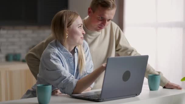 Ζευγάρι 30 γυναίκα και ο σύζυγος στην κουζίνα χρησιμοποιώντας φορητό υπολογιστή απολαμβάνουν e-shopping εξ αποστάσεως, σχεδιάζει μελλοντική μετεγκατάσταση επιλέξετε ανακαίνιση υπηρεσίες εταιρεία σε απευθείας σύνδεση. Ηλεκτρονικό εμπόριο εύκολη και άνετη δραστηριότητα από το σπίτι έννοια — Αρχείο Βίντεο