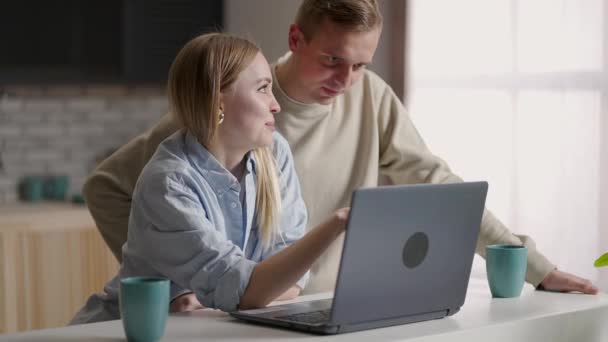 Pár 30-as évek felesége és férje a konyhában segítségével laptop élvezze az e-vásárlás távolról, tervezés jövőbeli áthelyezés választani felújítási cég szolgáltatások online. E-kereskedelem egyszerű és kényelmes tevékenység otthonról — Stock videók