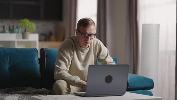 Серьезный молодой человек фрилансер, работающий на фрилансере из дома, печатающий электронную почту на ноутбуке, сфокусированный мужчина, использующий компьютер для учебы дома, сидящий на диване, мужчина, занятый на дистанционной интернет-работе — стоковое видео