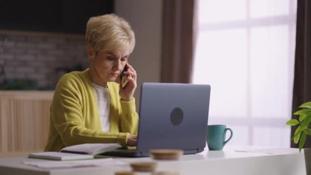 Ηλικιωμένη γυναίκα εργάζεται στο σπίτι, μιλώντας μέσω κινητού τηλεφώνου και πληκτρολογώντας σε φορητό υπολογιστή, γραφείο στο σπίτι και λειτουργία απομακρυσμένης εργασίας για ηλικιωμένους — Αρχείο Βίντεο