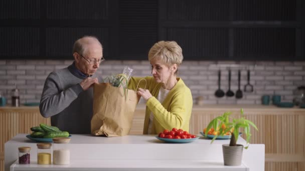 Gammal man och kvinna kontrollerar varor från leveransservice från matvaror, packa upp väska med mat i köket — Stockvideo
